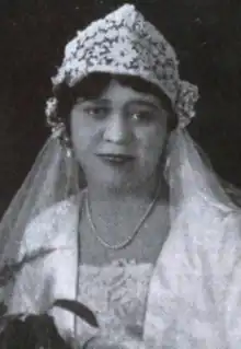 A young Black woman wearing a bridal ensemble, including a lace crown-shaped cap, a veil, a white jacket, and a lace dress