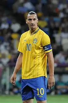 A photograph of a man with dark hair wearing a yellow football shirt, blue shorts and a dark blue captain's armband on his arm, the man is looking away from the camera.