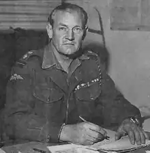 Black-and-white photograph of Churchill in uniform, sitting at a desk, and looking up at the camera while writing