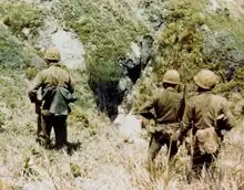 Three soldiers looking away from the camera toward a cave-like fissure.